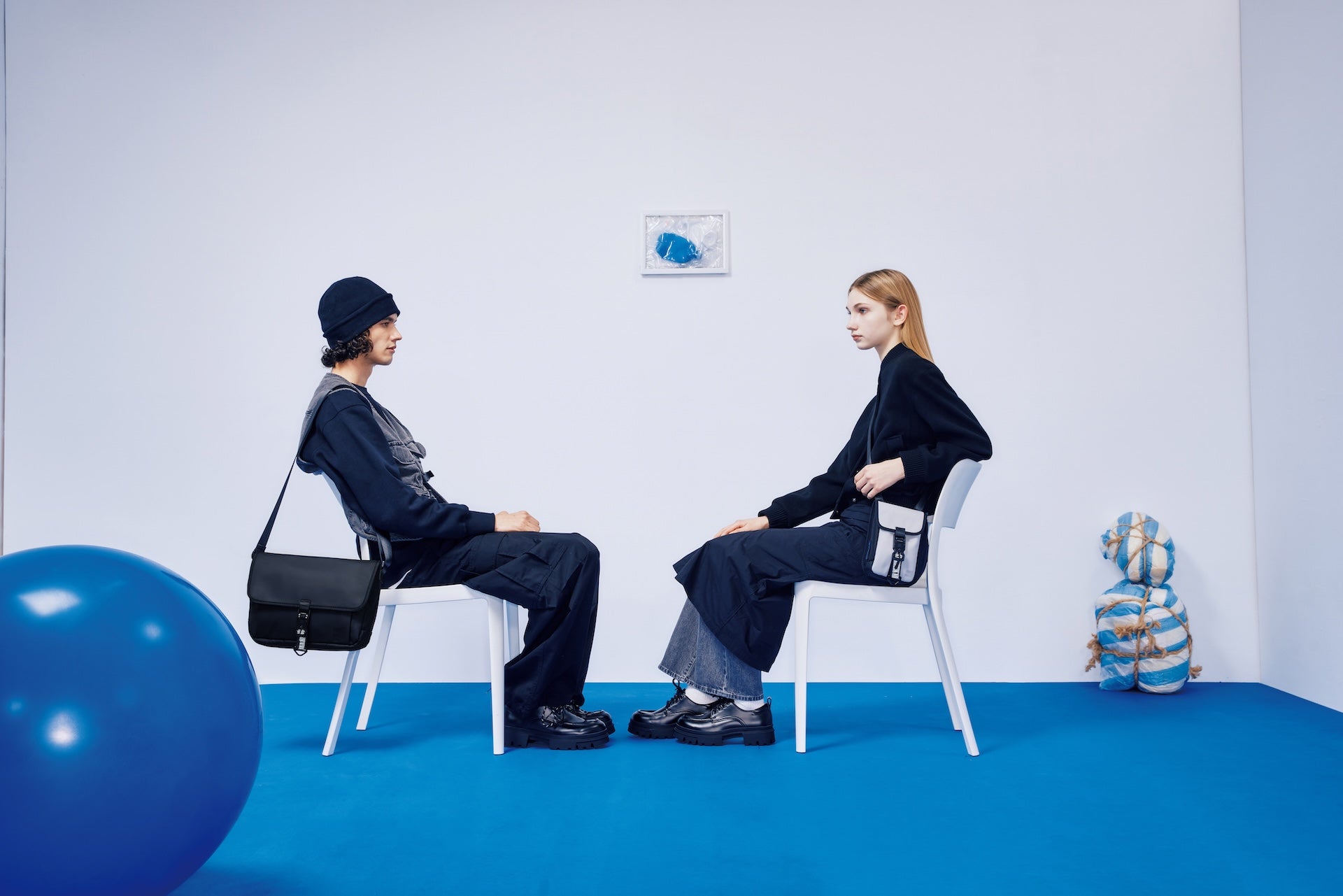 Two models sitting on chairs, each wearing sustainable bags, in a minimalist blue and white setting, highlighting eco-friendly fashion by WAVEIT.