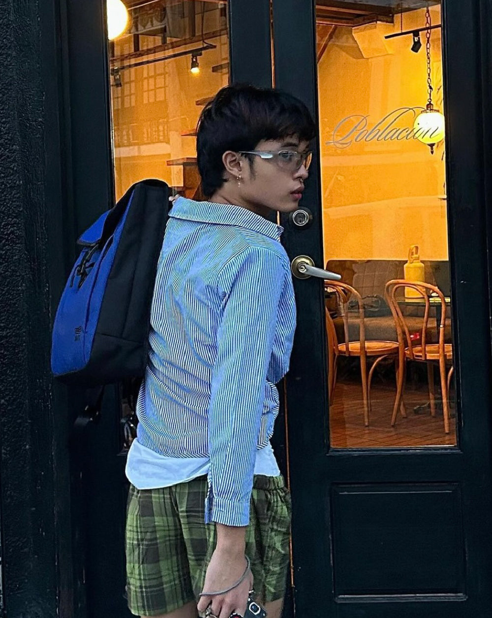 A young person with short hair and glasses stands in front of a glass door with a blue and black backpack slung over one shoulder. They are wearing a light blue striped shirt, white undershirt, and green plaid shorts. The interior of a cozy, warmly lit café with wooden chairs and tables can be seen through the door.