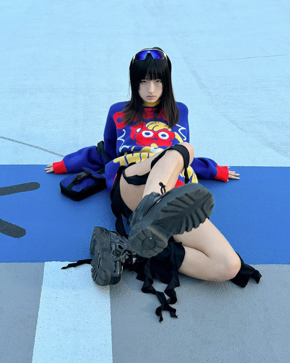 A young woman with straight black hair sits on the ground against a blue and gray background. She wears a vibrant sweater featuring a colorful character design, black boots, and reflective sunglasses resting on her head. Her legs are crossed, and she has a serious expression.