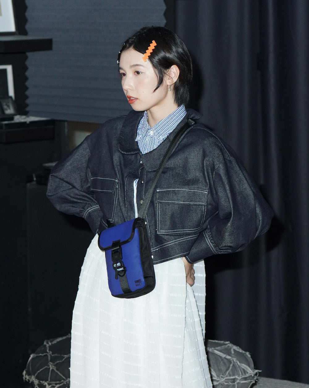 A young woman with short hair stands indoors wearing a dark denim jacket, a striped shirt, and a white skirt. She has two colorful hair clips in her hair and carries a blue and black crossbody bag. The background features dark curtains, and the text 'Sustainability In. Waste Out.' is overlaid on the image.