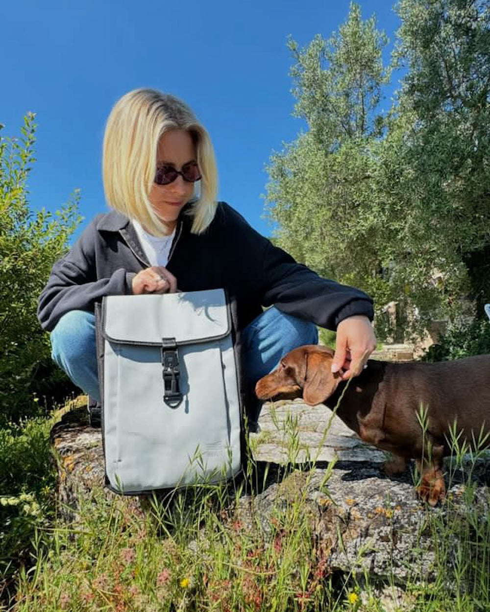 A woman with short blonde hair, wearing sunglasses, sits on a rock outside on a sunny day. She is petting a brown dachshund while holding a gray backpack. The background features lush green foliage and a clear blue sky.