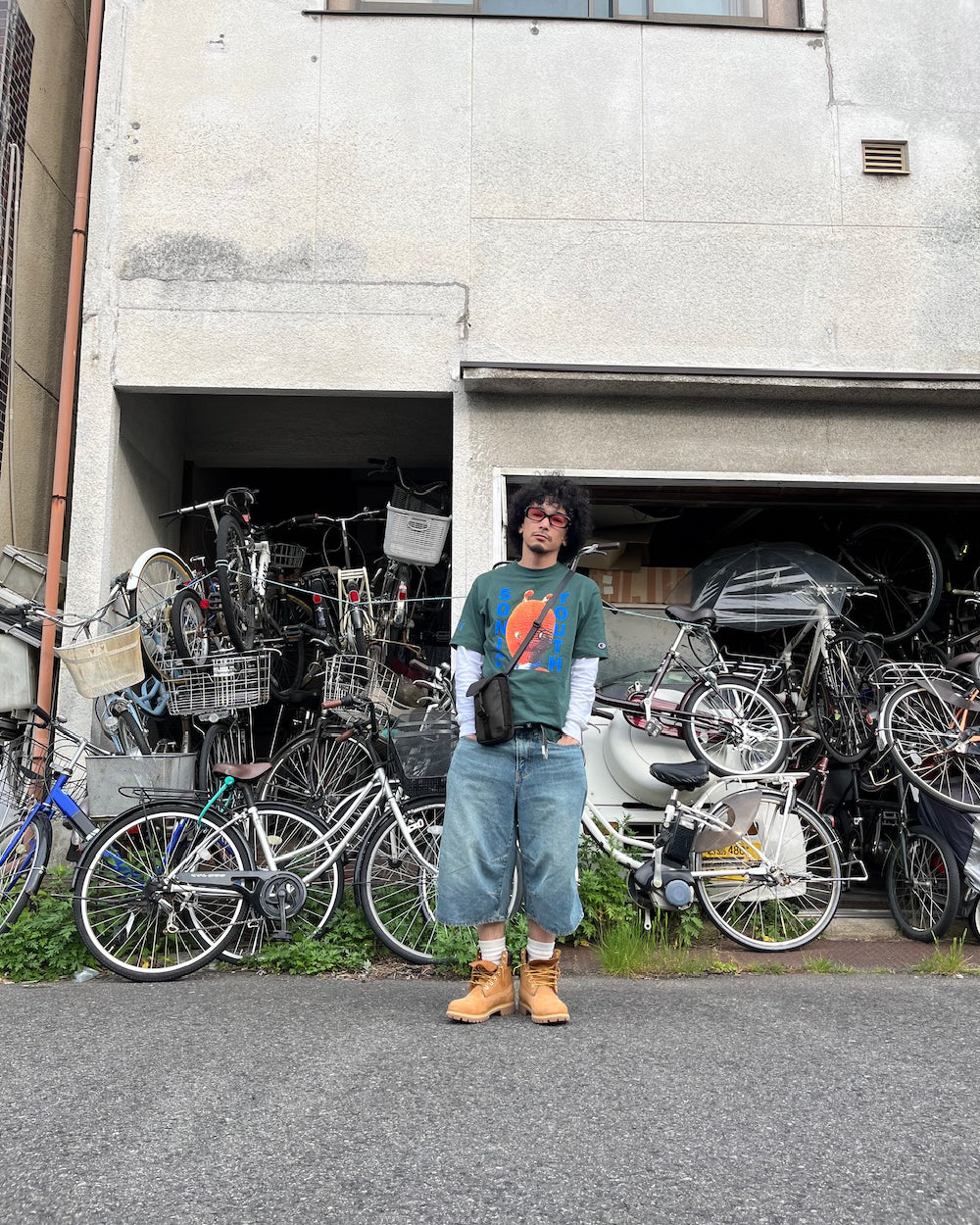 A man with curly hair and wearing red glasses stands in front of a building filled with stacked bicycles. He is dressed in a green graphic t-shirt, long sleeves underneath, denim shorts, and tan boots. The background features numerous bicycles in a storage area.