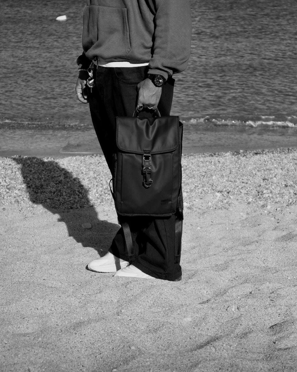 A black-and-white photo of a person standing on a sandy beach, holding a black backpack. The person is wearing a sweatshirt, loose-fitting pants, and white shoes. The ocean is visible in the background.