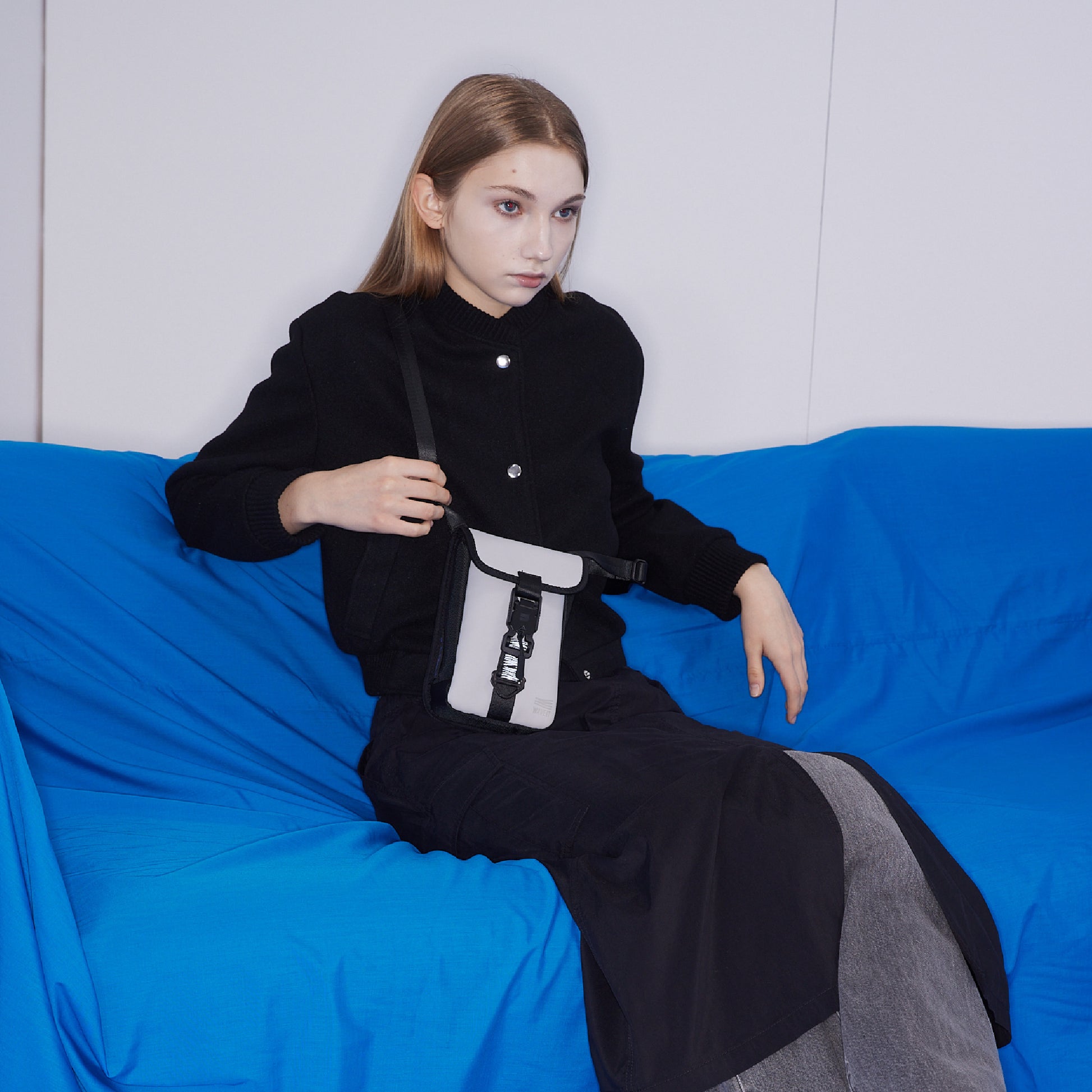 Model sitting on a blue sofa, wearing a black outfit with a white WAVEIT eco-friendly crossbody bag.