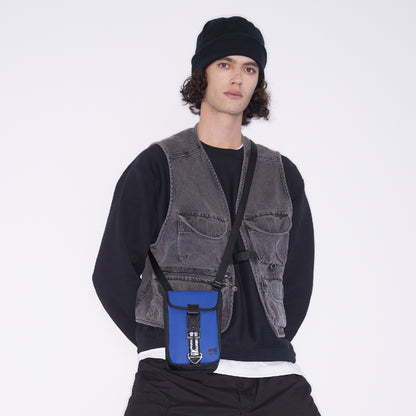 Model wearing WAVEIT blue eco-friendly crossbody bag with adjustable strap, styled with a casual outfit including a beanie and utility vest.